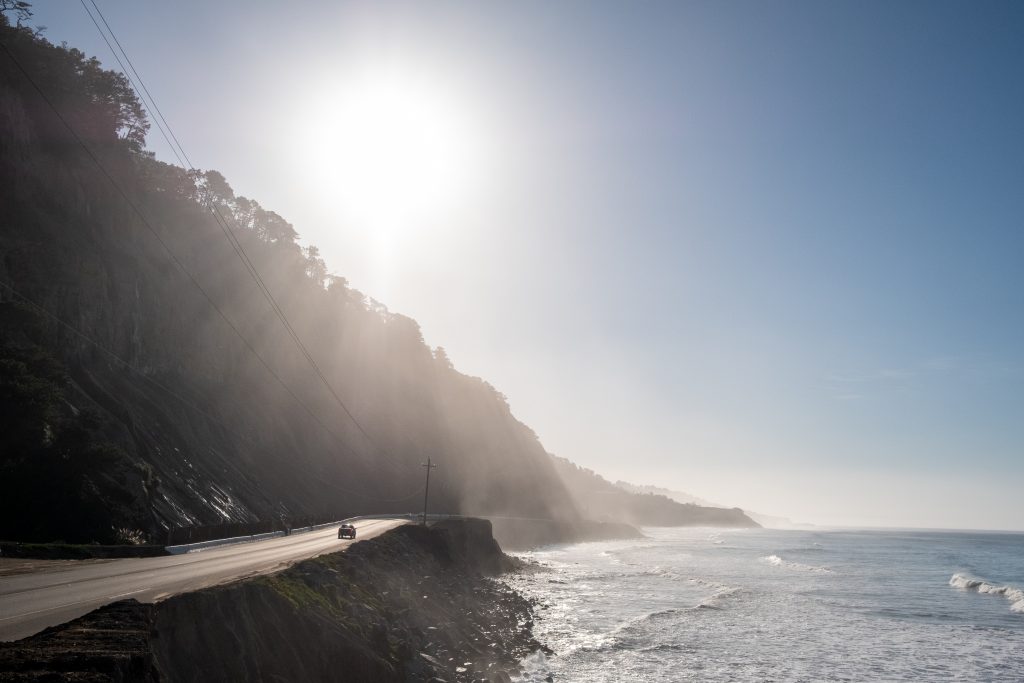 Le soleil brille au dessus d’une crête donnant sur le bord de mer. Plus loin, une voiture est visible.